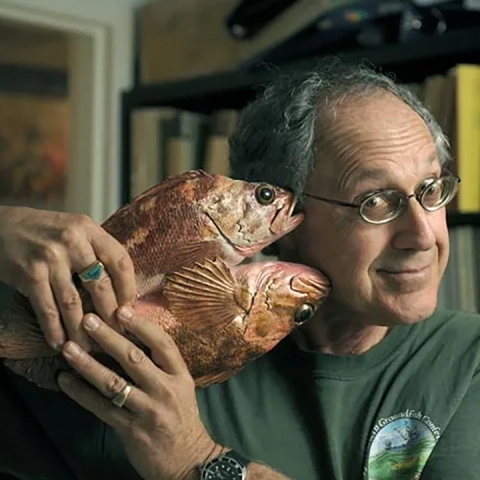 Milton Love sits in front of a bookshelf in a t-shirt, holding a statue of two fish next to his head