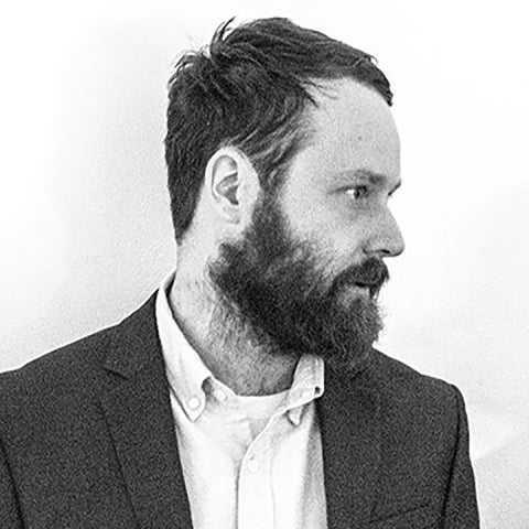 Alex Lukas, in a black & white, stands in front of a wall, looking away to the right, wearing a suit jacket 