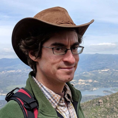 Marc Mayes stands at a vista point overlooking a town, wearing a wide-brimmed brown hat and jacket