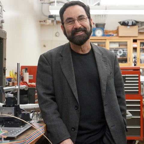 Philip Lubin stands in a lab and wears a dark blazer over a black t-shirt