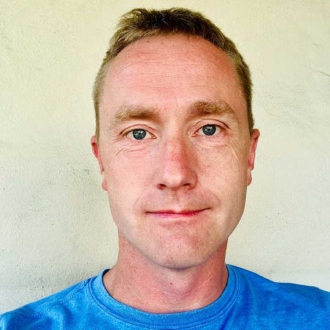 David Weld stands against a wall wearing a blue t-shirt