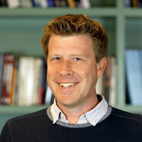 Andy Howell wears a sweater over a collared shirt, standing in front of a bookshelf