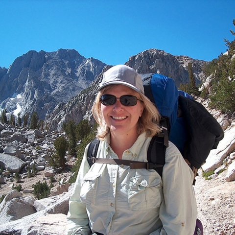 Cherie Briggs stands on a mountain, wearing a hat, sunglasses, and backpack.