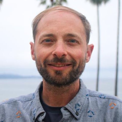 Gavin McDonald wears a collared shirt near some palm trees and a body of water