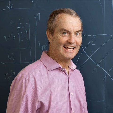 Joseph Polchinski wears a collared shirt and stands in front of a chalkboard