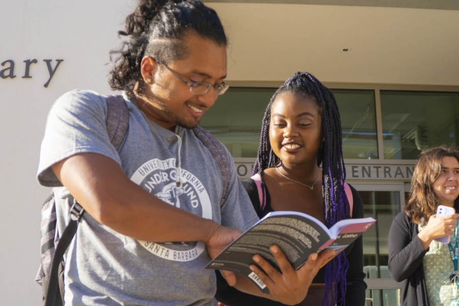 Man with brown hair and skin holds book as woman with black hair and braids looks at him
